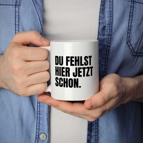 Person hält Tasse mit Spruch „DU FEHLST HIER JETZT SCHON.“ in beiden Händen