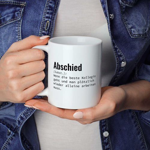 Person hält Tasse mit Spruch „Abschied [Subst.]: Wenn die beste Kollegin geht und man plötzlich wieder alleine arbeiten muss.“ in beiden Händen