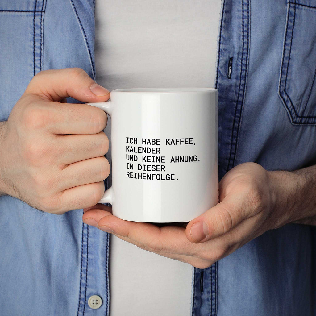 Person hält weiße Tasse mit dem Spruch „Ich habe Kaffee, Kalender und keine Ahnung“ in beiden Händen