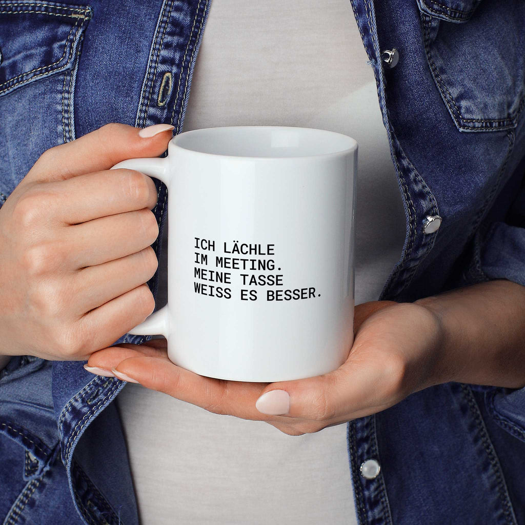 Person hält weiße Tasse mit dem Spruch „Ich lächle im Meeting. Meine Tasse weiß es besser“ in beiden Händen