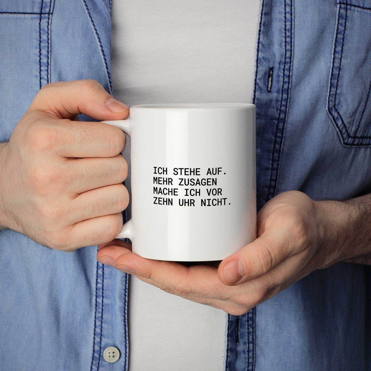 Person hält weiße Tasse mit dem Spruch „Mehr Zusagen mache ich vor zehn Uhr nicht“ in beiden Händen