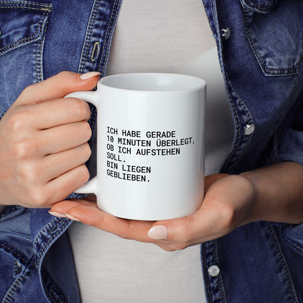 Weiße Tasse „Ich habe gerade 10 Minuten überlegt, ob ich aufstehen soll....“ in beiden Händen gehalten
