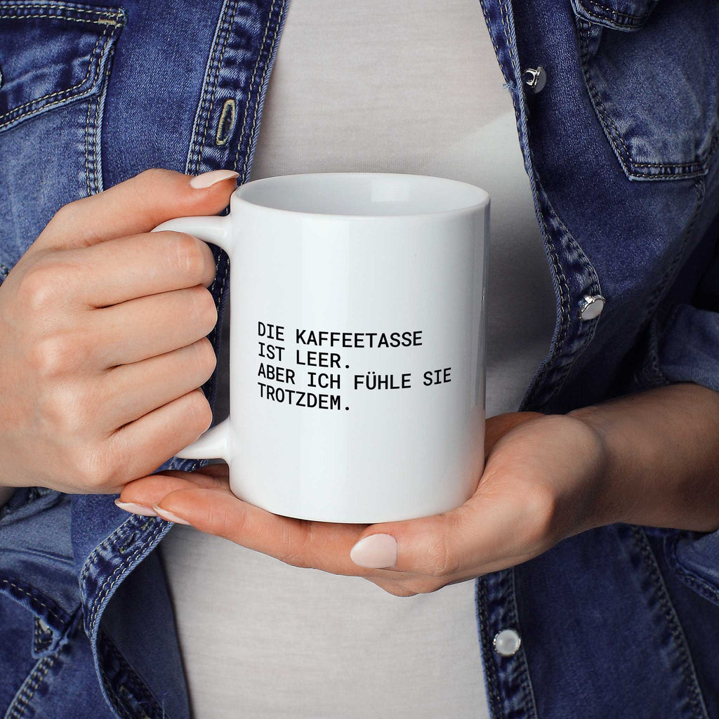 Weiße Tasse „Die Kaffeetasse ist leer – aber ich fühle sie trotzdem.“ in beiden Händen gehalten