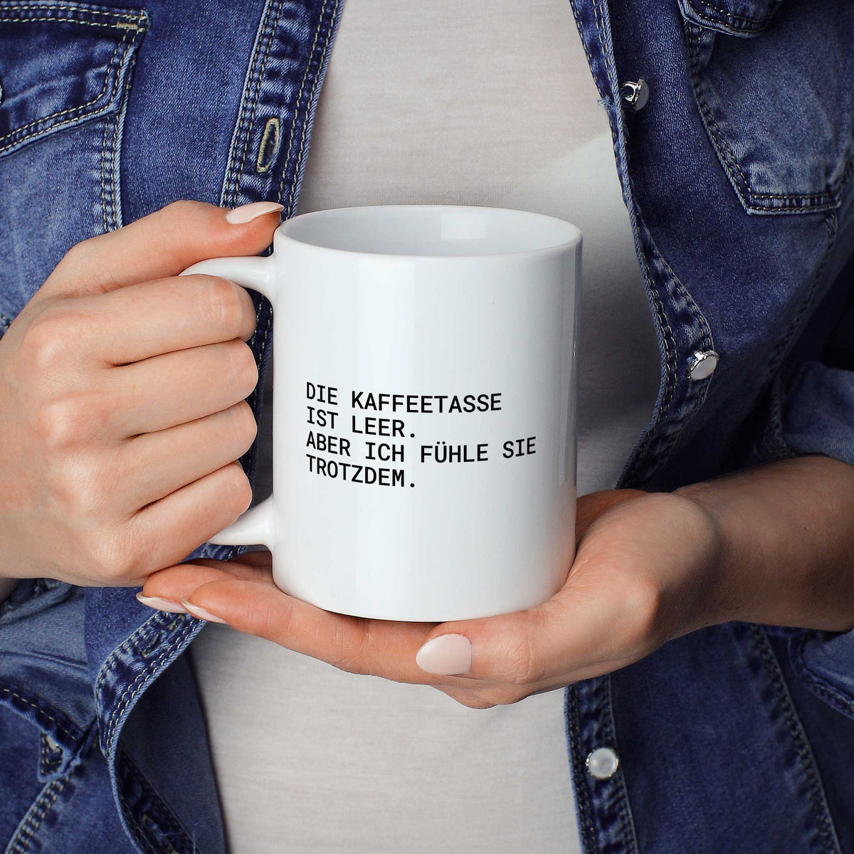 Weiße Tasse „Die Kaffeetasse ist leer – aber ich fühle sie trotzdem.“ in beiden Händen gehalten