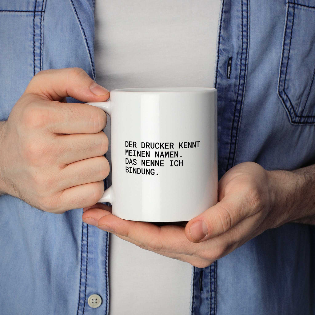 Weiße Tasse „Der Drucker kennt meinen Namen. Das nenne ich Bindung.“ in beiden Händen gehalten