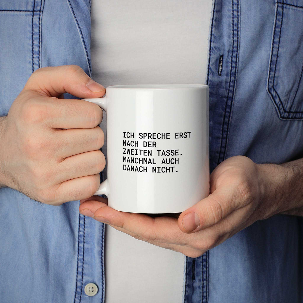 Weiße Tasse „Ich spreche erst nach der zweiten Tasse. Manchmal auch danach...“ in beiden Händen gehalten