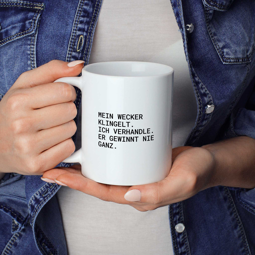 Weiße Tasse „Mein Wecker klingelt. Ich verhandle. Er gewinnt nie ganz.“ in beiden Händen gehalten