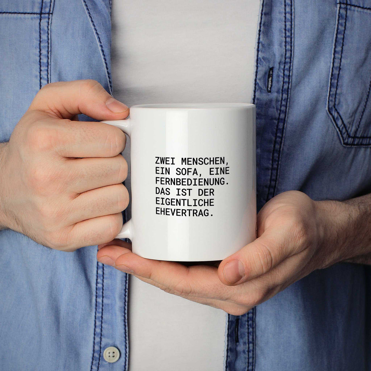 Weiße Tasse „Zwei Menschen, ein Sofa, eine Fernbedienung – das ist der...“ in beiden Händen gehalten