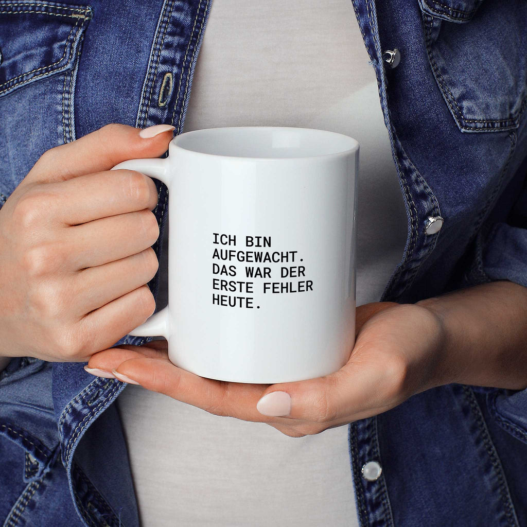 Weiße Tasse „Ich bin aufgewacht – das war der erste Fehler heute.“ in beiden Händen gehalten