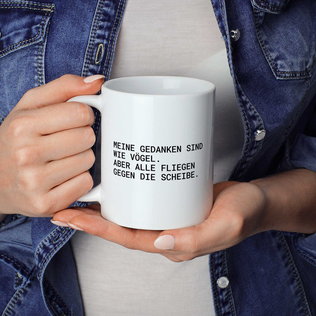Weiße Tasse „Meine Gedanken sind wie Vögel – aber alle fliegen gegen die...“ in beiden Händen gehalten