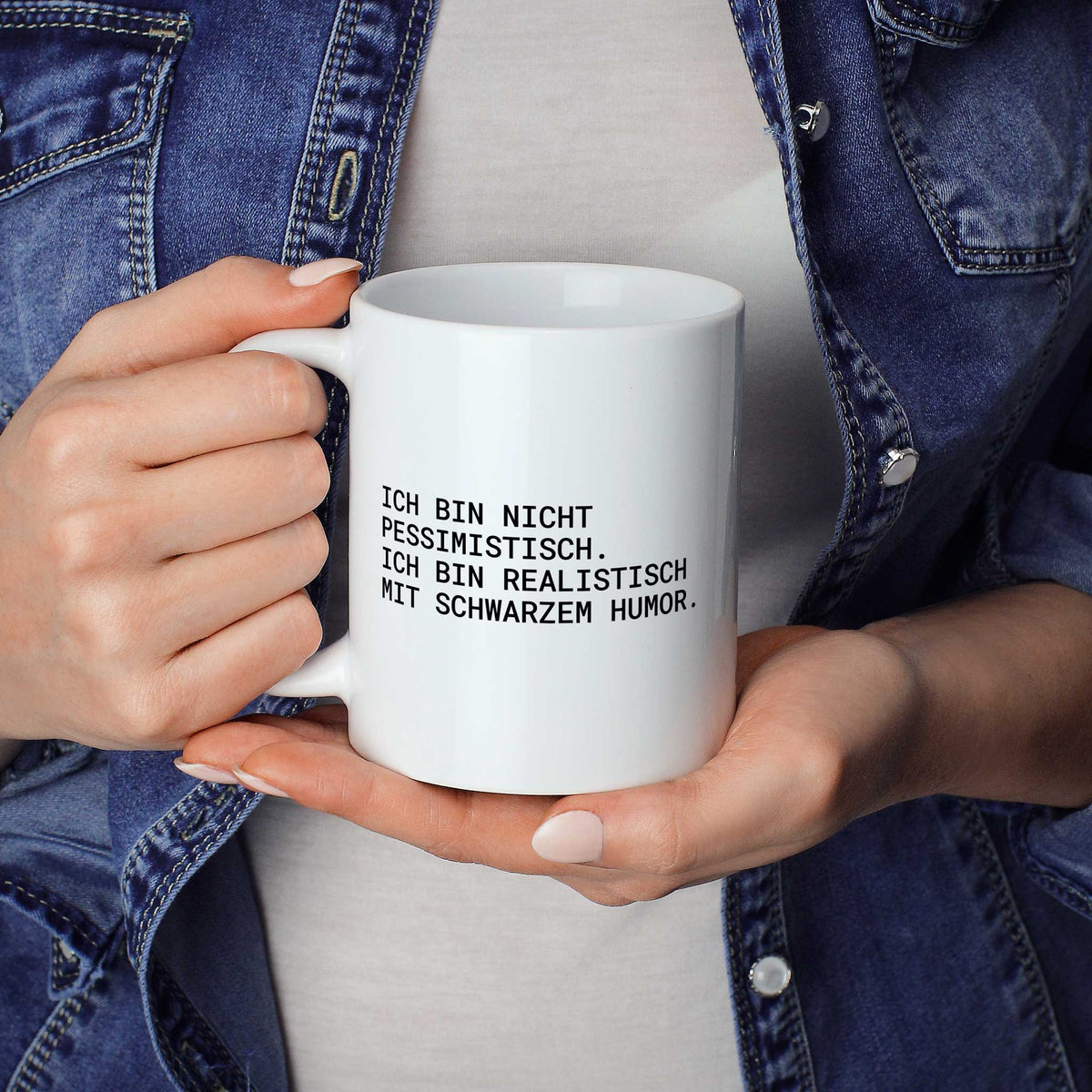 Weiße Tasse „Ich bin nicht pessimistisch – ich bin realistisch mit...“ in beiden Händen gehalten