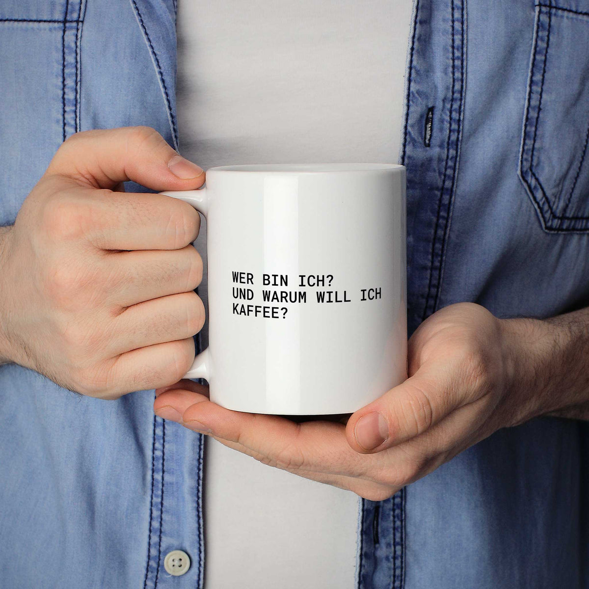 Weiße Tasse „Wer bin ich? – und warum will ich Kaffee?“ in beiden Händen gehalten