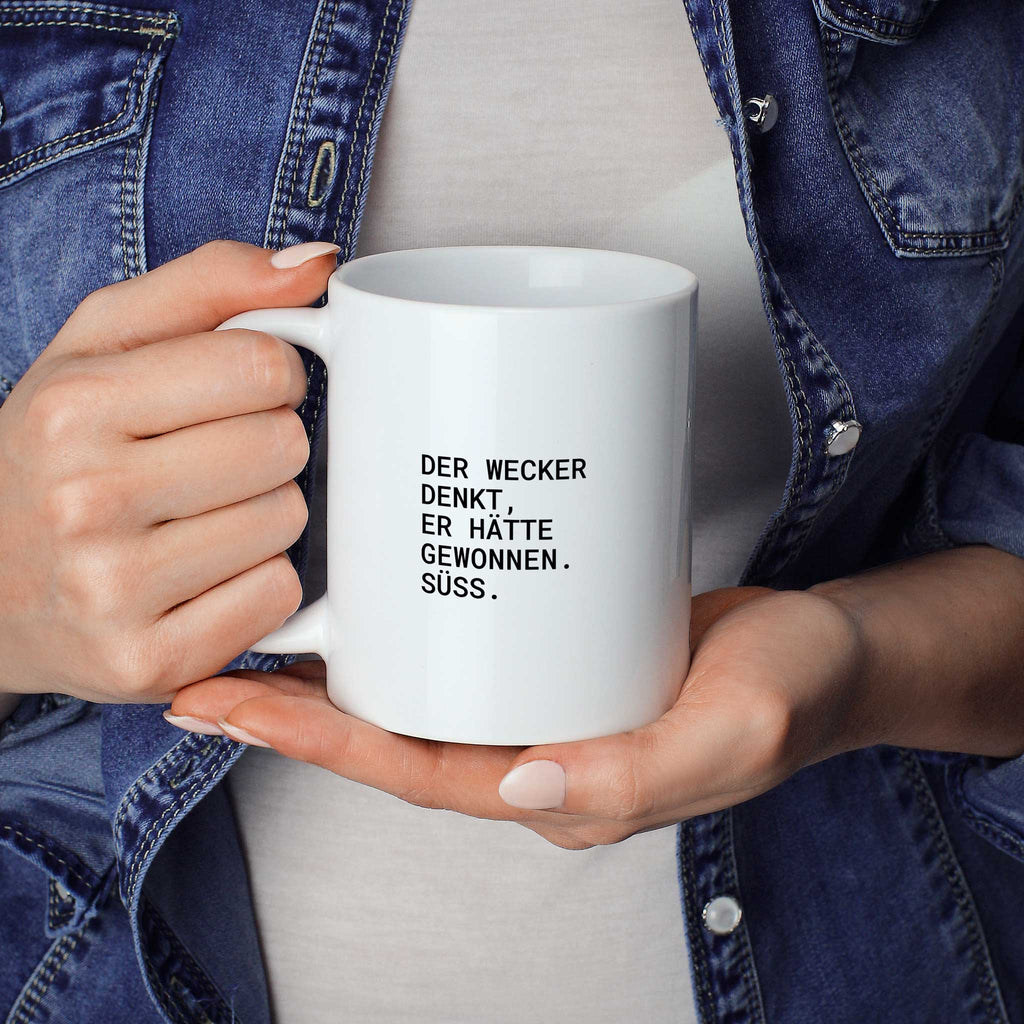 Person hält weiße Tasse mit dem Spruch „Der Wecker denkt, er hätte gewonnen. Süß.“ in beiden Händen