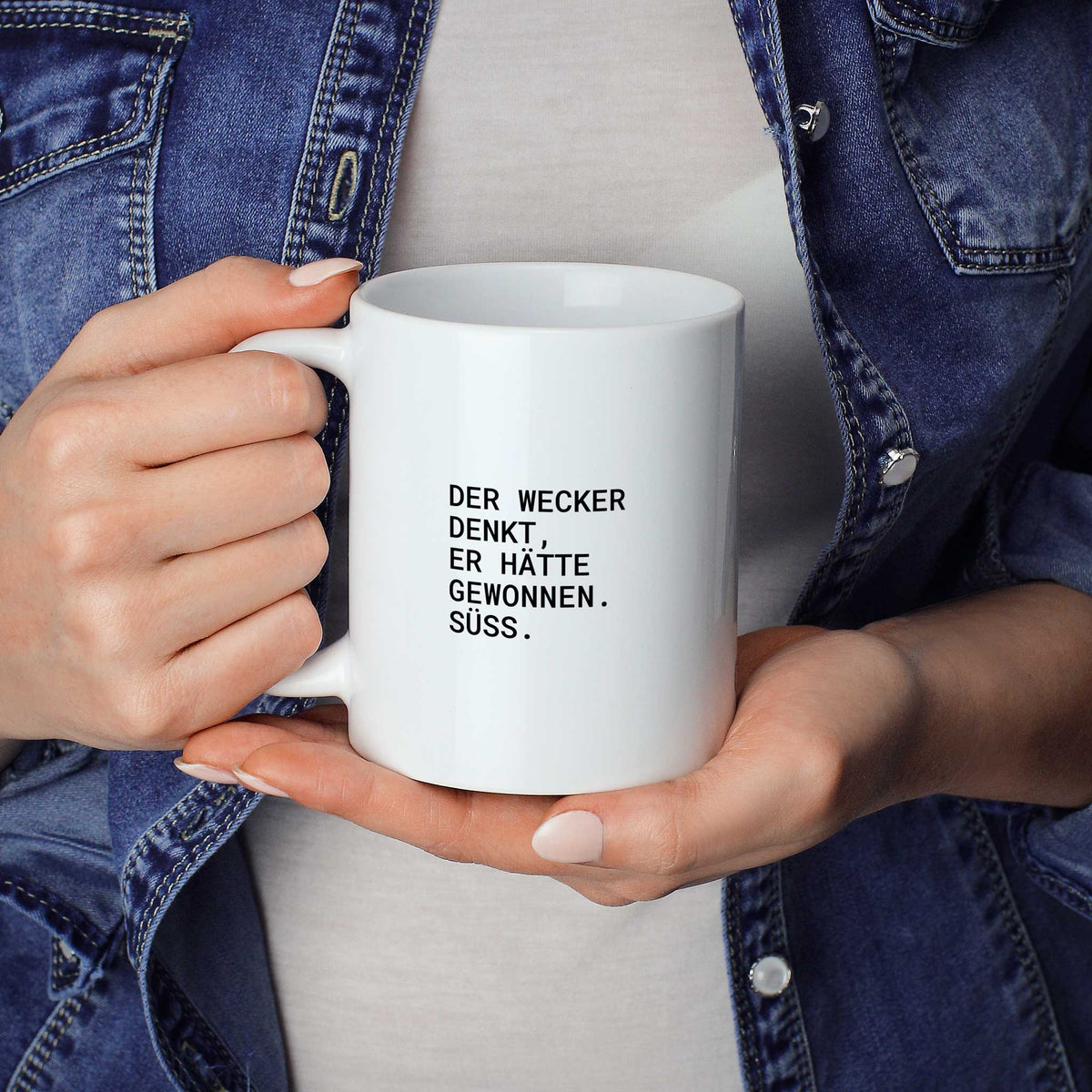 Person hält weiße Tasse mit dem Spruch „Der Wecker denkt, er hätte gewonnen. Süß.“ in beiden Händen