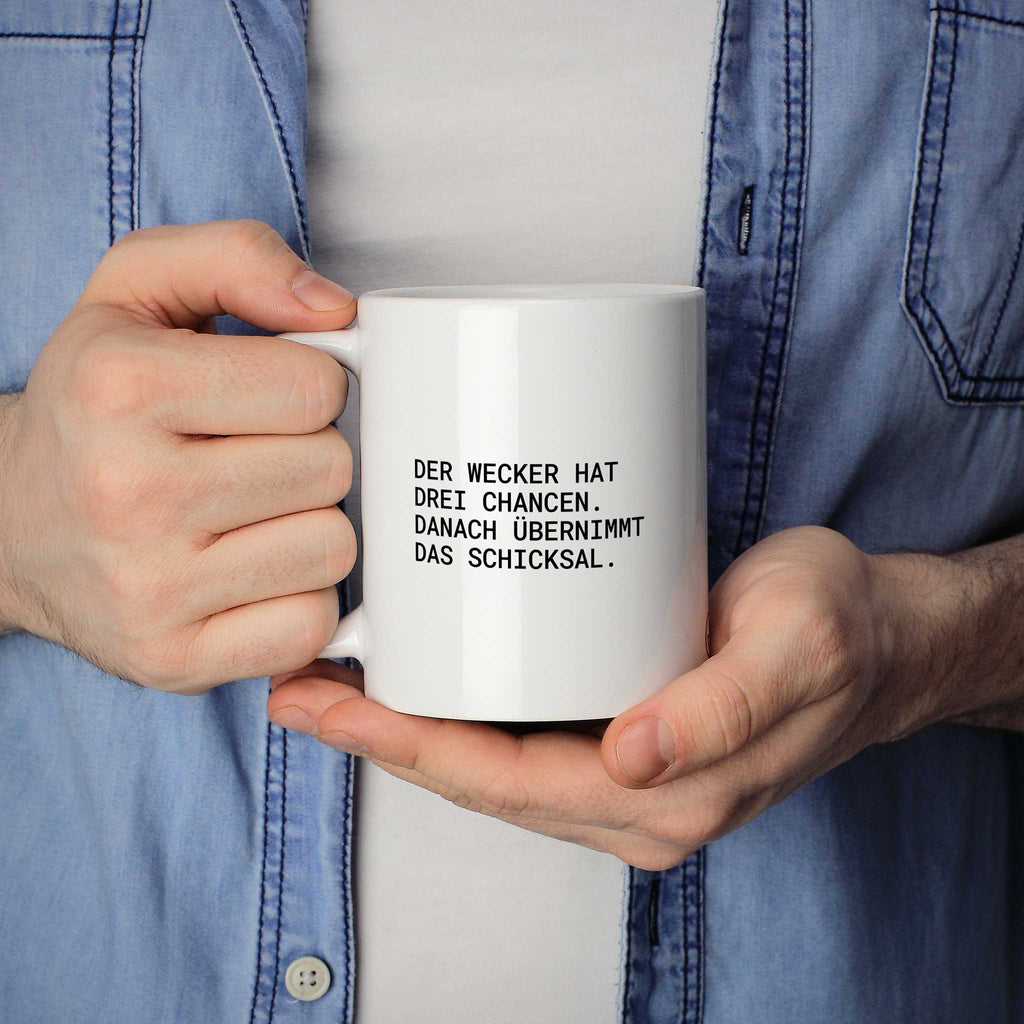 Person hält weiße Tasse mit dem Spruch „Der Wecker hat drei Chancen“ in beiden Händen