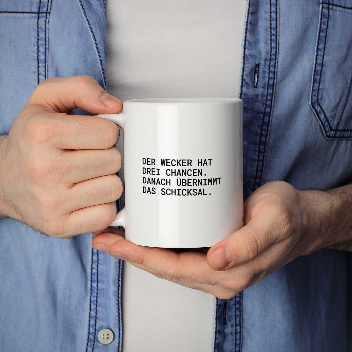 Person hält weiße Tasse mit dem Spruch „Der Wecker hat drei Chancen“ in beiden Händen
