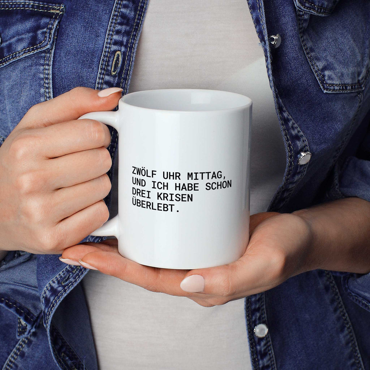 Person hält weiße Tasse mit dem Spruch „Zwölf Uhr Mittag, und ich habe schon drei Krisen“ in beiden Händen